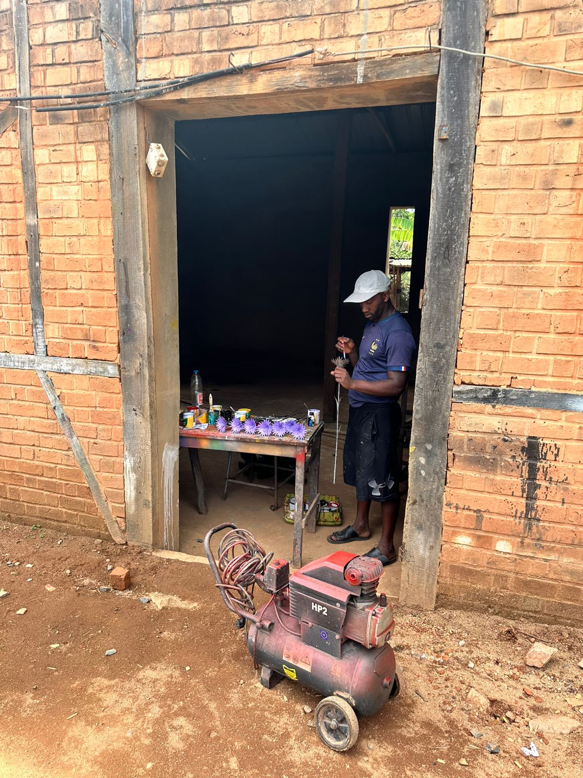 A man painting small game pieces at a workbench in the doorway of a brick workshop, with paint tins beside him and an air compressor on the ground outside.