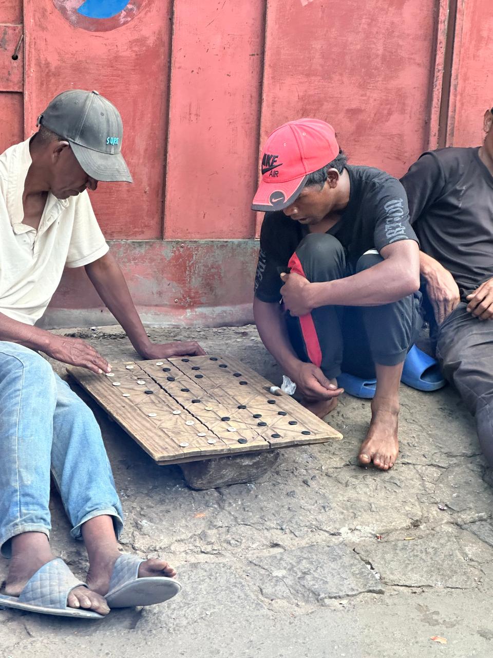 Two men crouched over a hand-cut wooden board on a pavement, playing with small black and white discs, while a third man watches from beside them.