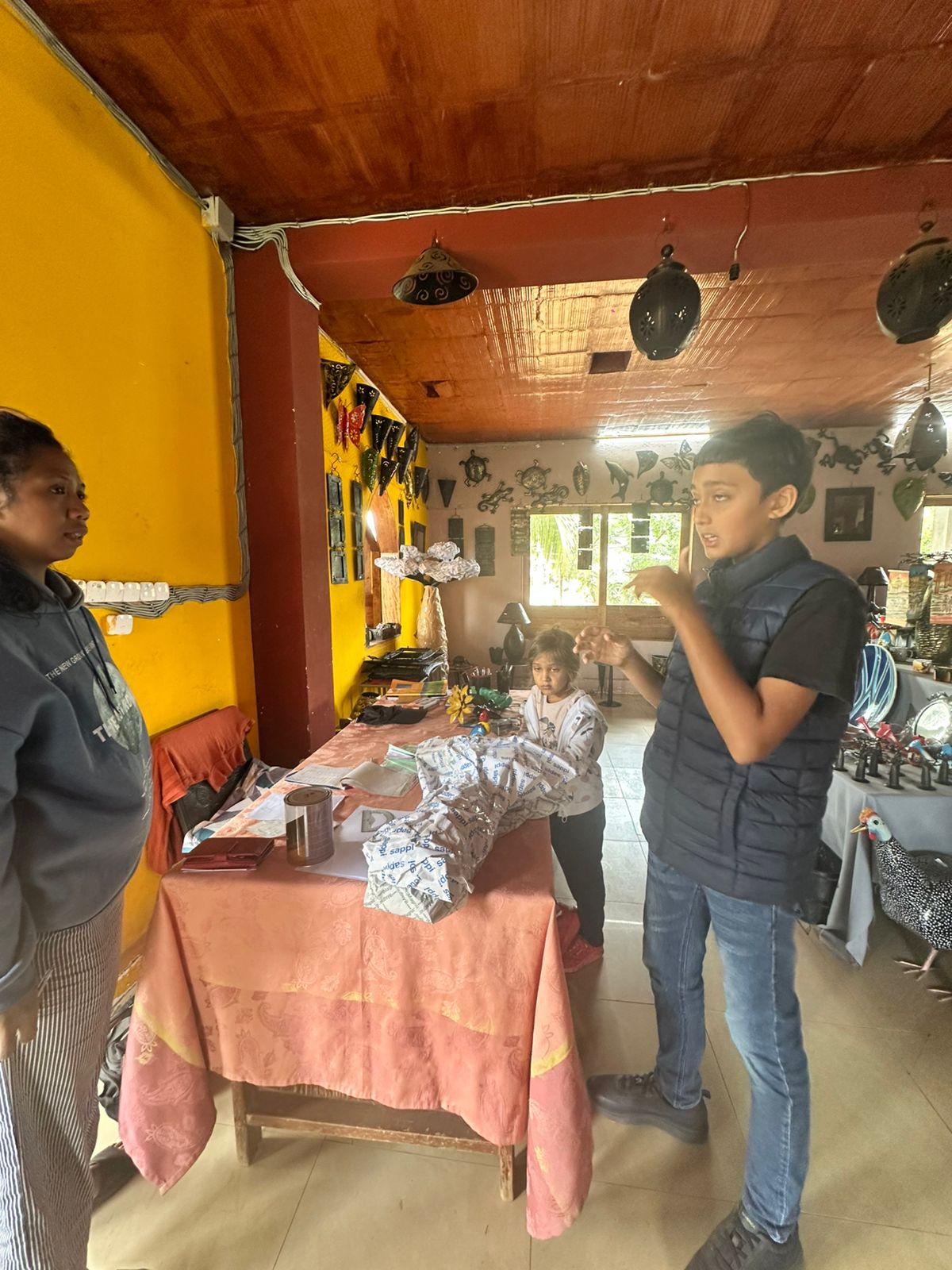 Two children and an adult talking across a cloth-covered workbench in a Malagasy craft workshop hung with cut-metal ornaments.